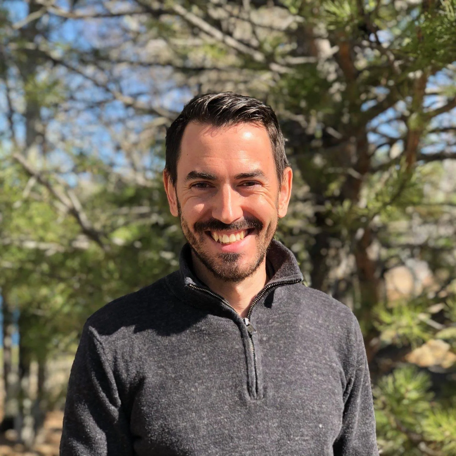 A smiling man with dark hair and a beard wearing a dark gray zip-up jacket stands outdoors in front of a tree with green leaves and a blue sky.