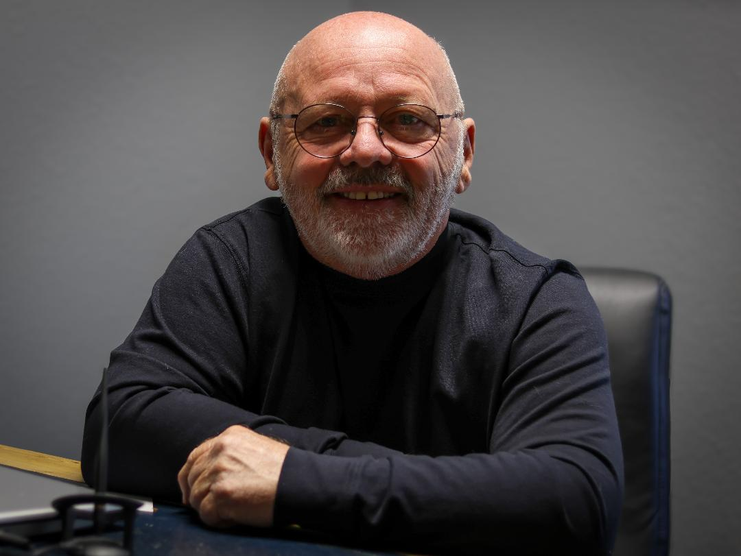 An older man with a beard and glasses sitting at a table, smiling at the camera, wearing a black shirt, against a plain gray background.