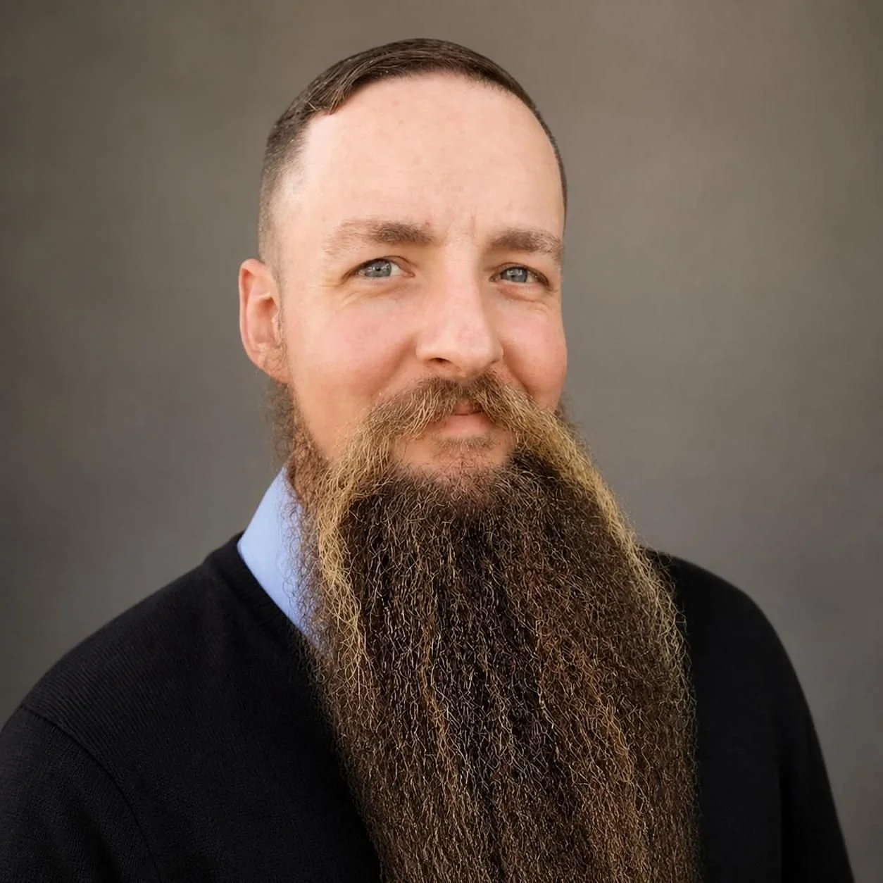 A man with a long beard and short hair taking a selfie outdoors in front of a green hedge.