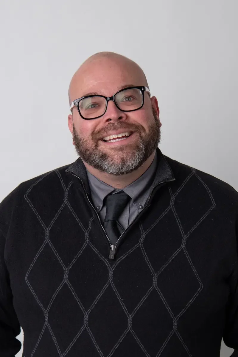 A smiling man with a beard and glasses, wearing a black sweater over a gray shirt and gray tie, against a plain white background.
