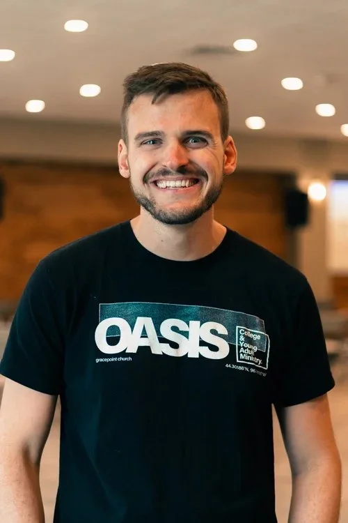 A smiling young man with short brown hair and facial hair standing indoors, wearing a black t-shirt with the word 'OASIS' and additional text.