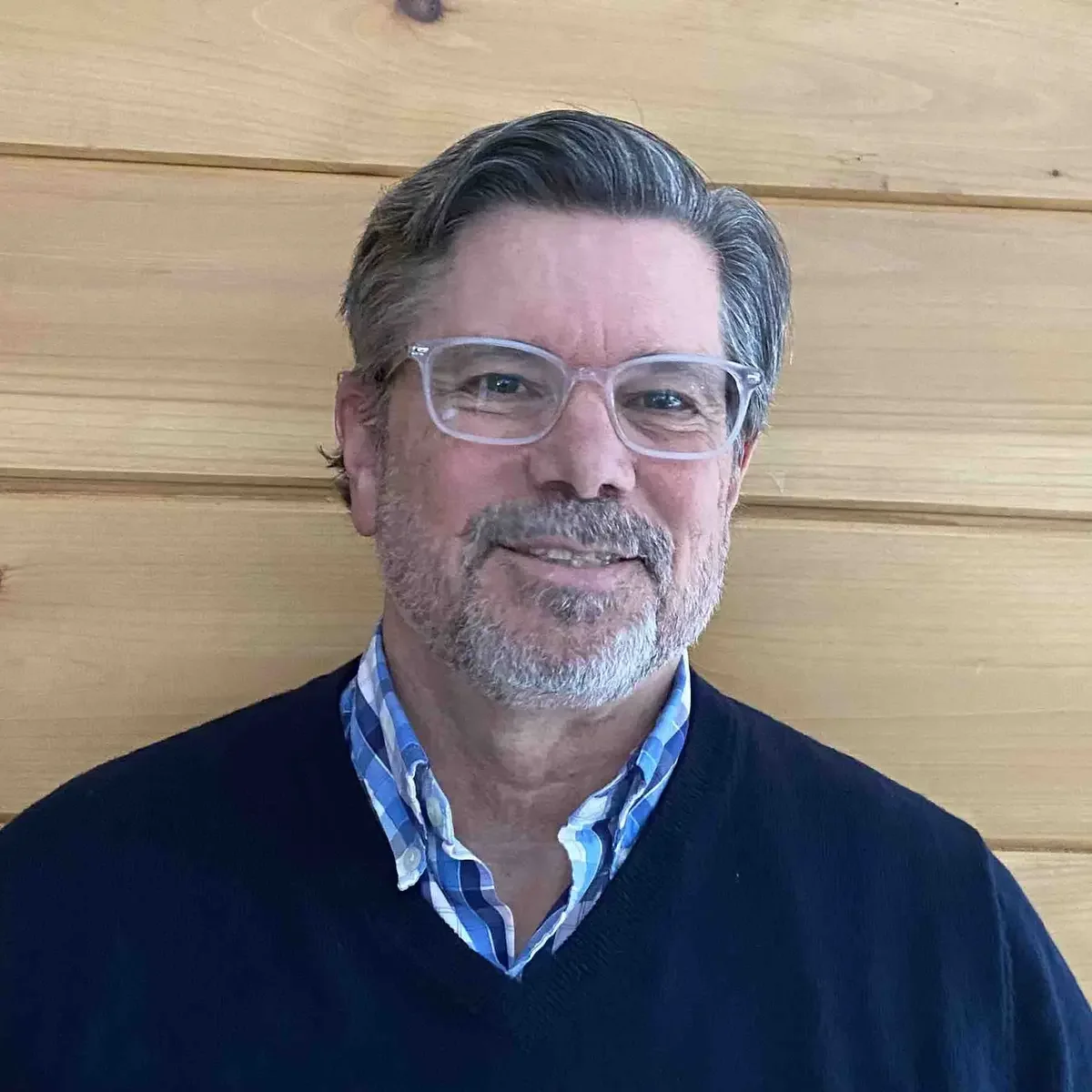 A man with gray hair and a beard, wearing clear glasses, a checkered blue shirt, and a black sweater, standing in front of a wooden panel wall.