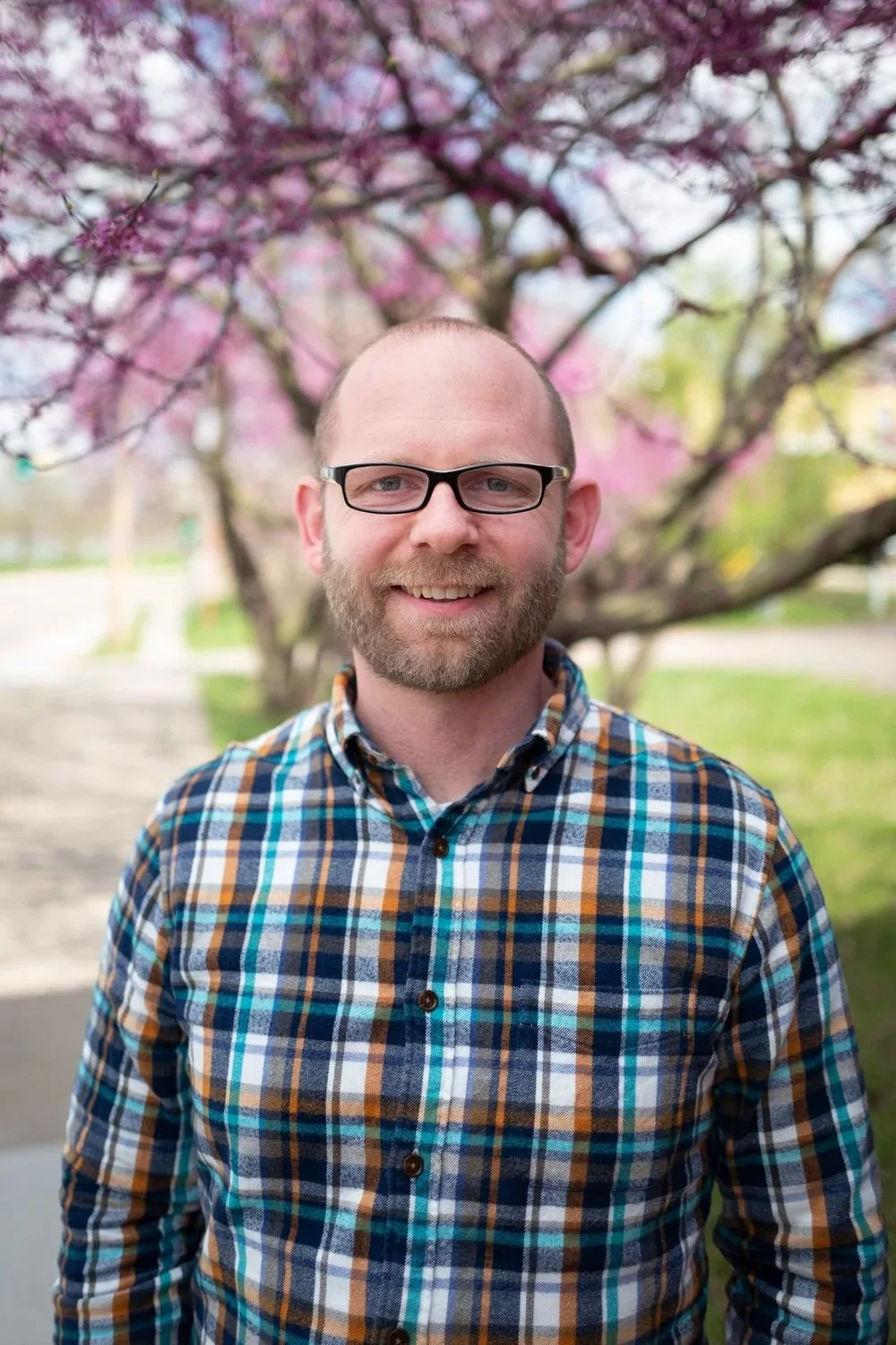 A man with glasses and a beard standing outdoors in front of a pink flowering tree, smiling at the camera.