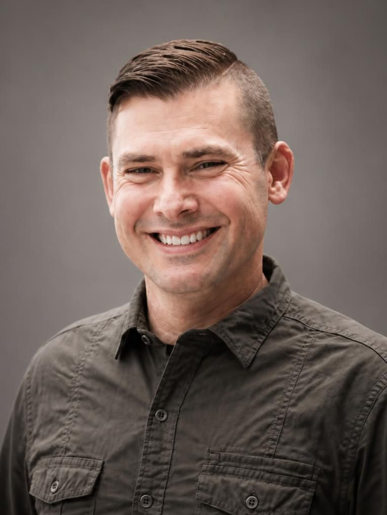 A smiling young man with short hair, wearing a button-up shirt, standing outdoors with a blurred natural background.
