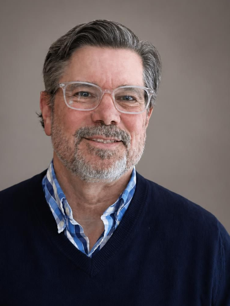 A man with gray hair and a beard, wearing clear glasses, a checkered blue shirt, and a black sweater, standing in front of a wooden panel wall.