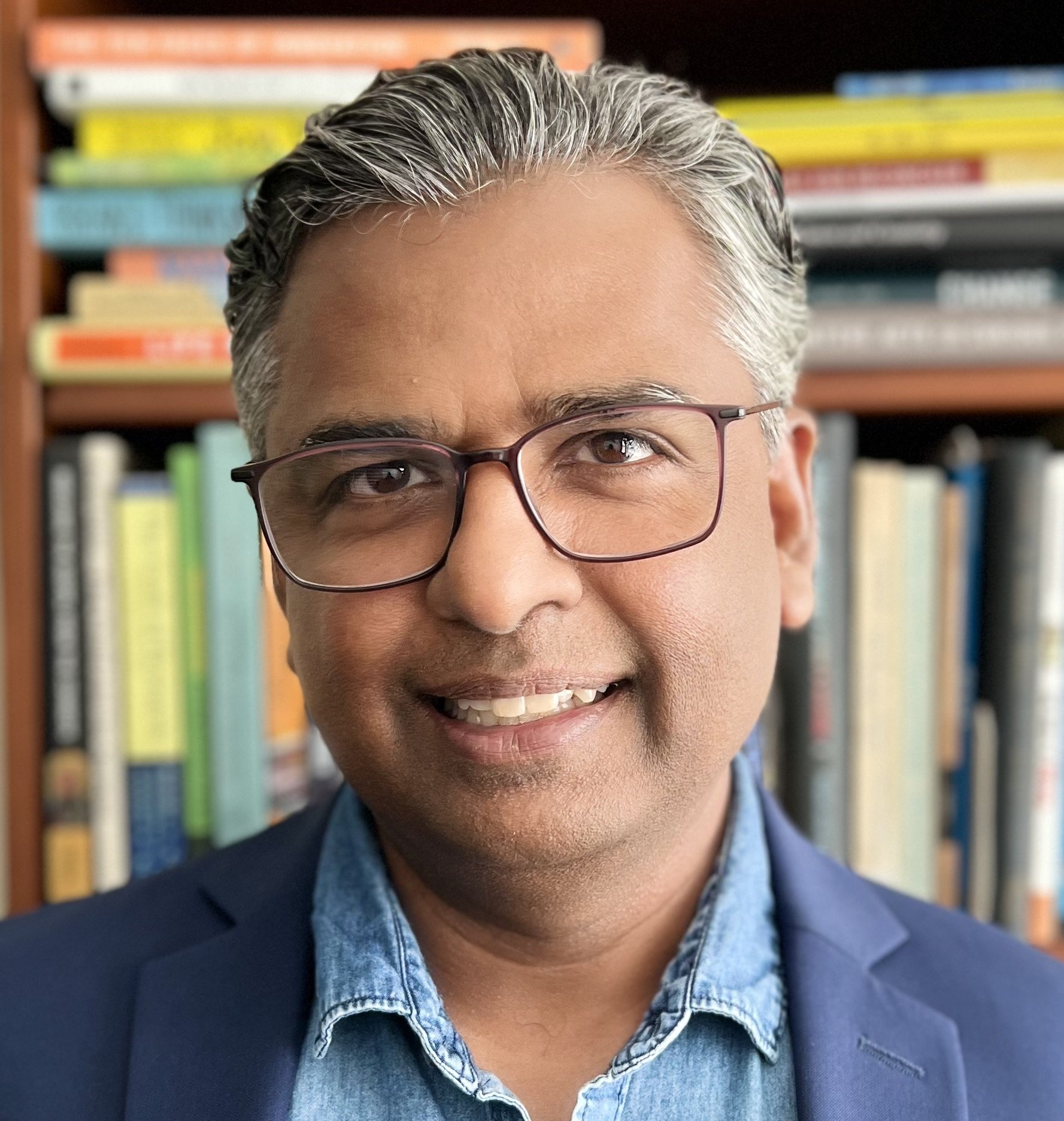Close-up of a smiling person with glasses, gray hair, wearing a denim shirt and blazer, with bookshelves in the background.