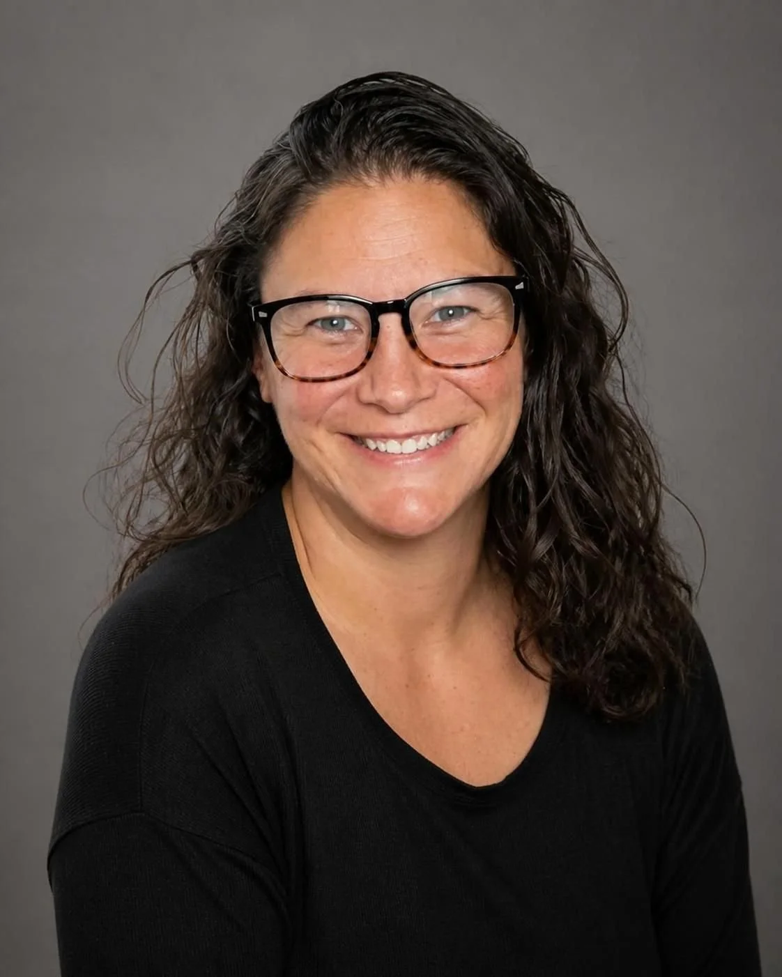 A woman with curly brown hair, wearing glasses and a black shirt, smiling and waving at the camera indoors.
