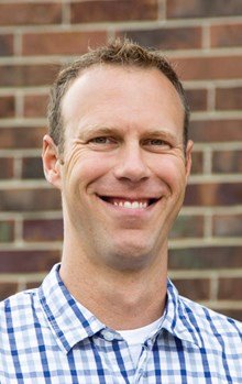 Portrait of a smiling man with short curly hair, wearing a blue checkered shirt, standing in front of a brick wall.