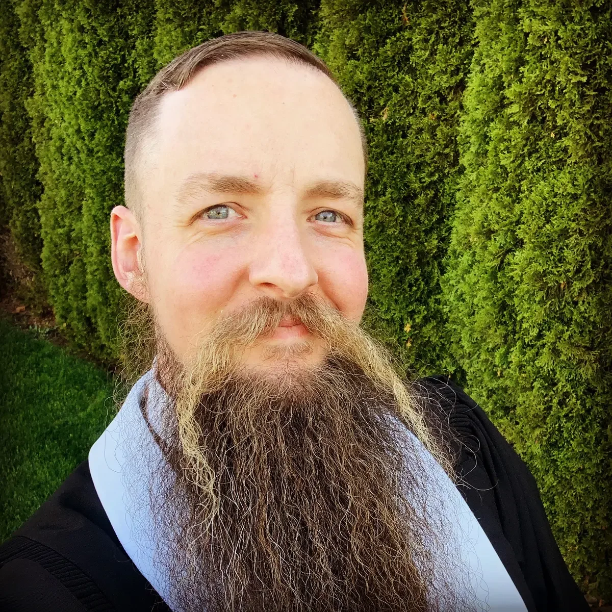 A man with a long beard and short hair taking a selfie outdoors in front of a green hedge.
