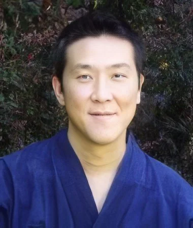 A man with black hair in a blue martial arts gi standing outdoors in front of trees.