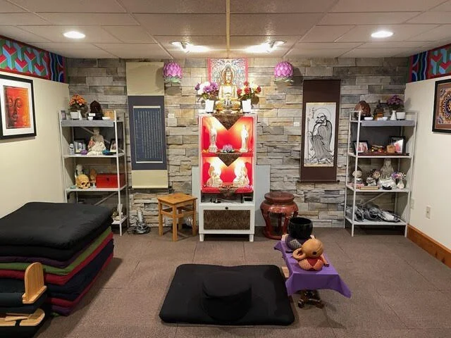 Room with a central altar featuring a Buddha statue, surrounded by shelves with decorative items and framed photos, a black cushion on the floor, and other traditional and modern decor elements.