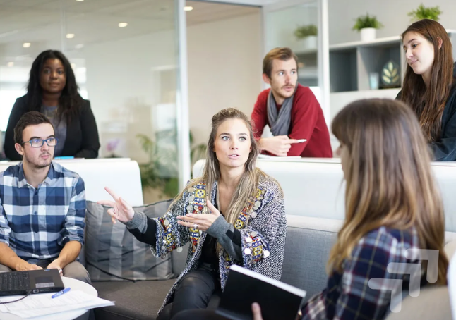 A diverse group of six young adults engaged in a discussion in a modern office setting with plants and glass walls.