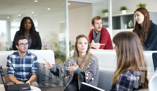 Group of diverse young adults having a discussion in a modern office, with some sitting and others standing, and a woman in the center speaking.
