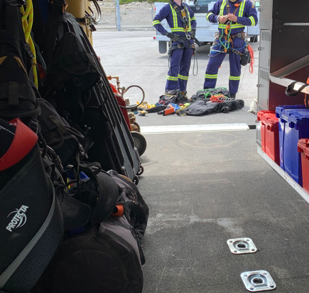 Interior of a rescue truck with safety gear and equipment, including helmets and harnesses. Two workers in safety uniforms and helmets are standing outside, organizing climbing or rescue gear.