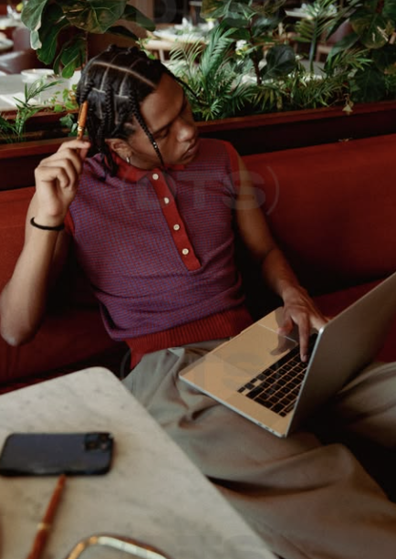 Person with braided hair sitting on a red booth seat, using a laptop with a plant and greenery in the background.