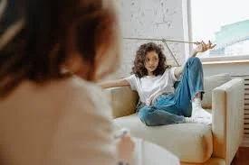 A young girl sitting on a couch playfully pointing a stick or wand at a woman who is sitting opposite her, out of focus in the foreground.