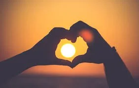 Two people form a heart shape with their hands around the setting sun during sunset