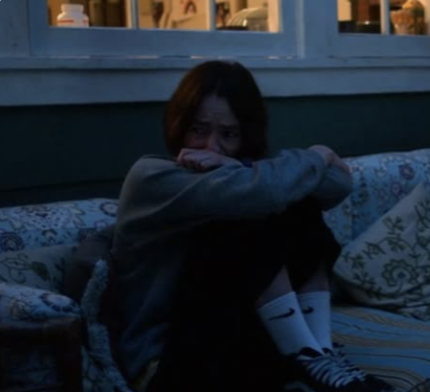 A person sitting on a patterned couch with arms around knees, illuminated by faint blue light, in a dimly lit room.