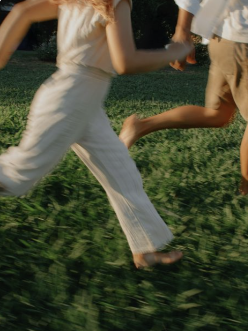 Two people running barefoot through a grassy field during sunset.