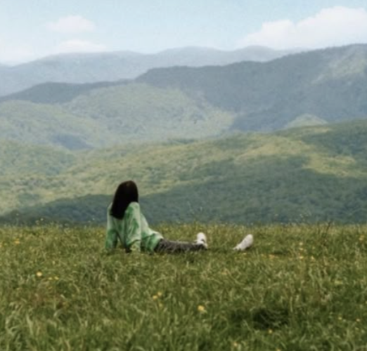 Person sitting in a grassy field with mountains in the background.