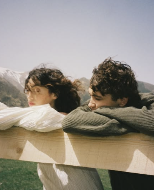 Two people resting on a wooden railing outdoors with mountains in the background.