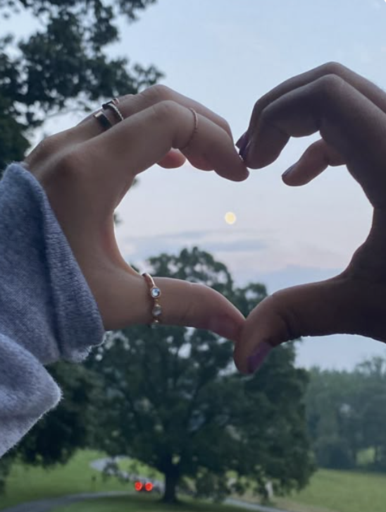 Two hands forming a heart shape with the moon in the background during dusk or dawn in a park.