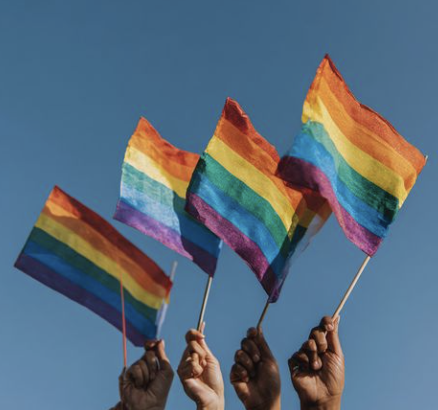 Four hands holding rainbow pride flags against a blue sky.