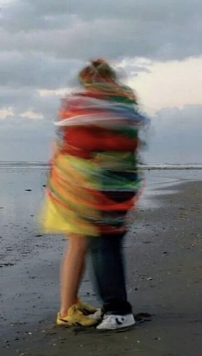 Two people hugging on a beach with a cloudy sky, wrapped in colorful blankets or clothing.