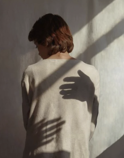 A woman with short brown hair facing away from the camera, standing against a plain wall, with shadows of hands cast on her back and shoulder.
