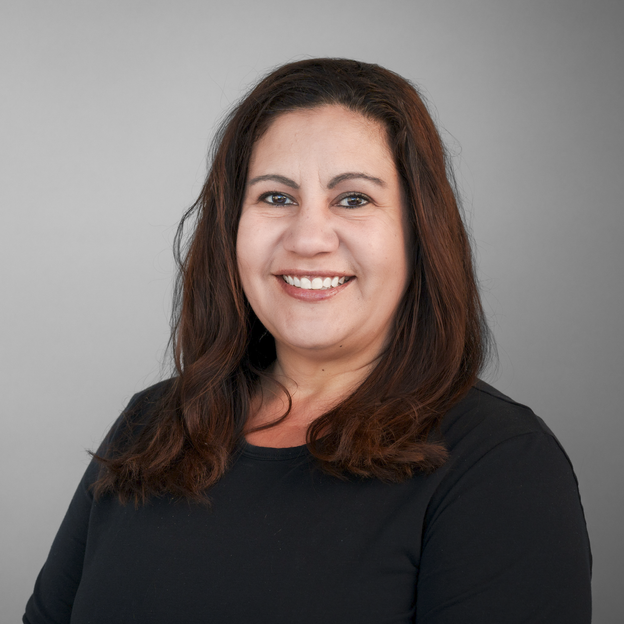 A dental hygienist with dark, wavy hair smiling at the camera, wearing a black scrub top.