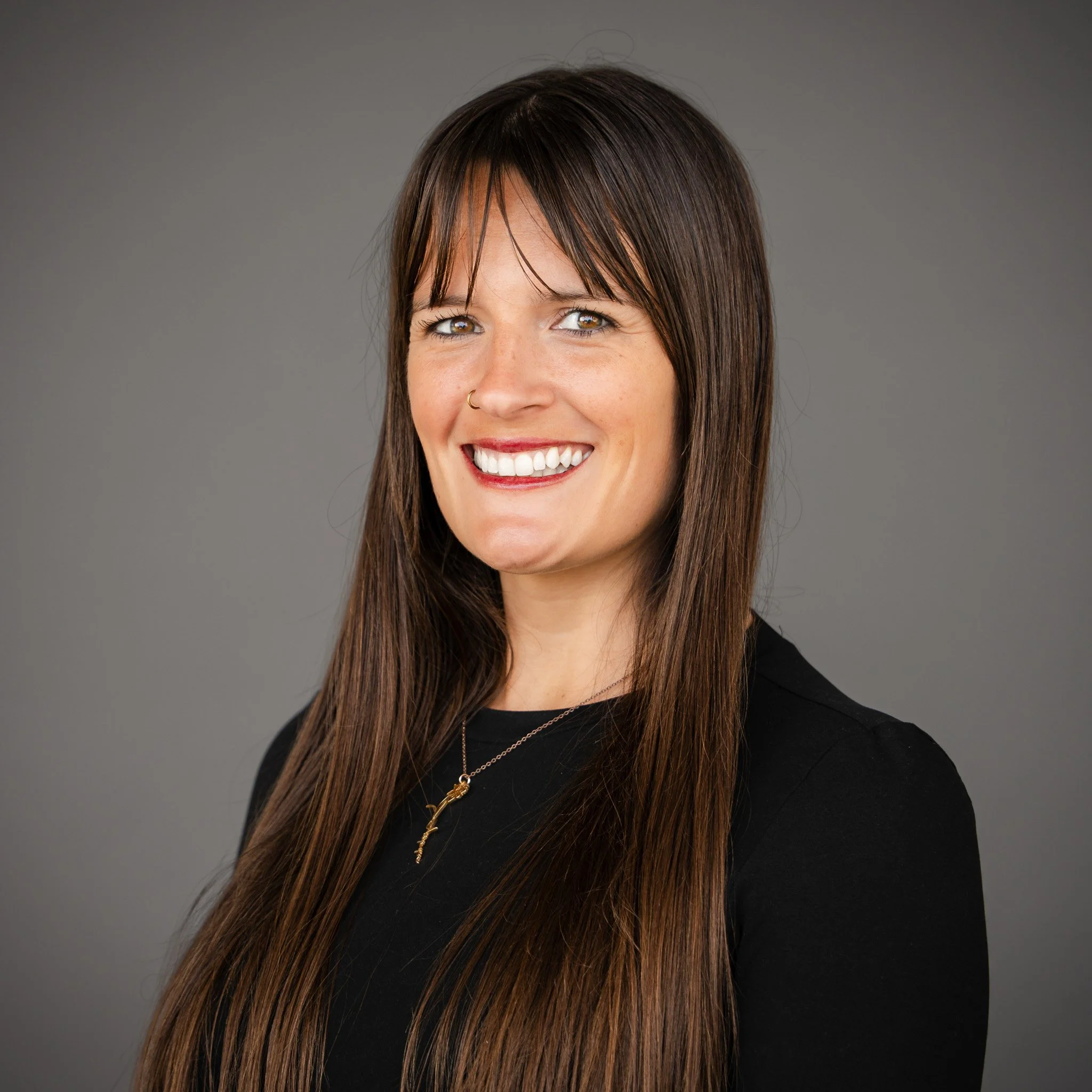 A young woman with long brown hair, smiling, standing outdoors in front of green trees and a sidewalk, wearing a dark teal blouse and a delicate gold necklace.