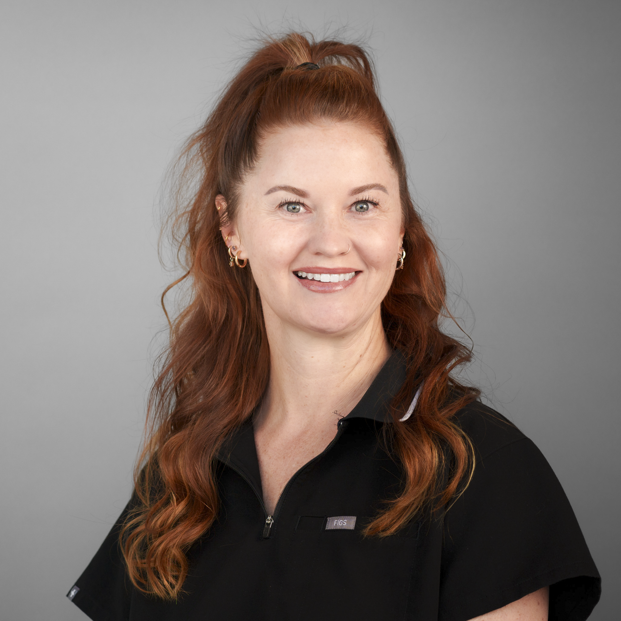 Smiling person with red hair in a bun, wearing a black collared shirt, standing in front of a stone wall.