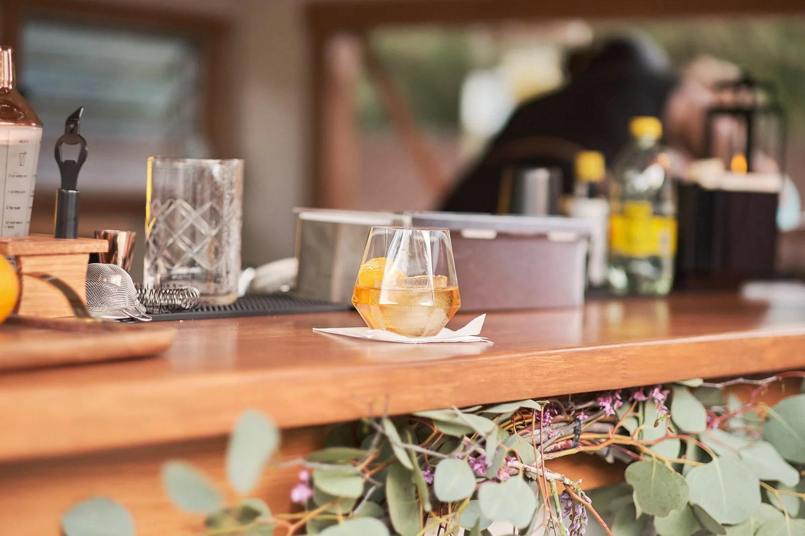 A cocktail sitting on a napkin on a wooden bar counter, with various bar tools and bottles in the background. Towing Temptations Mobile Bar Trailer