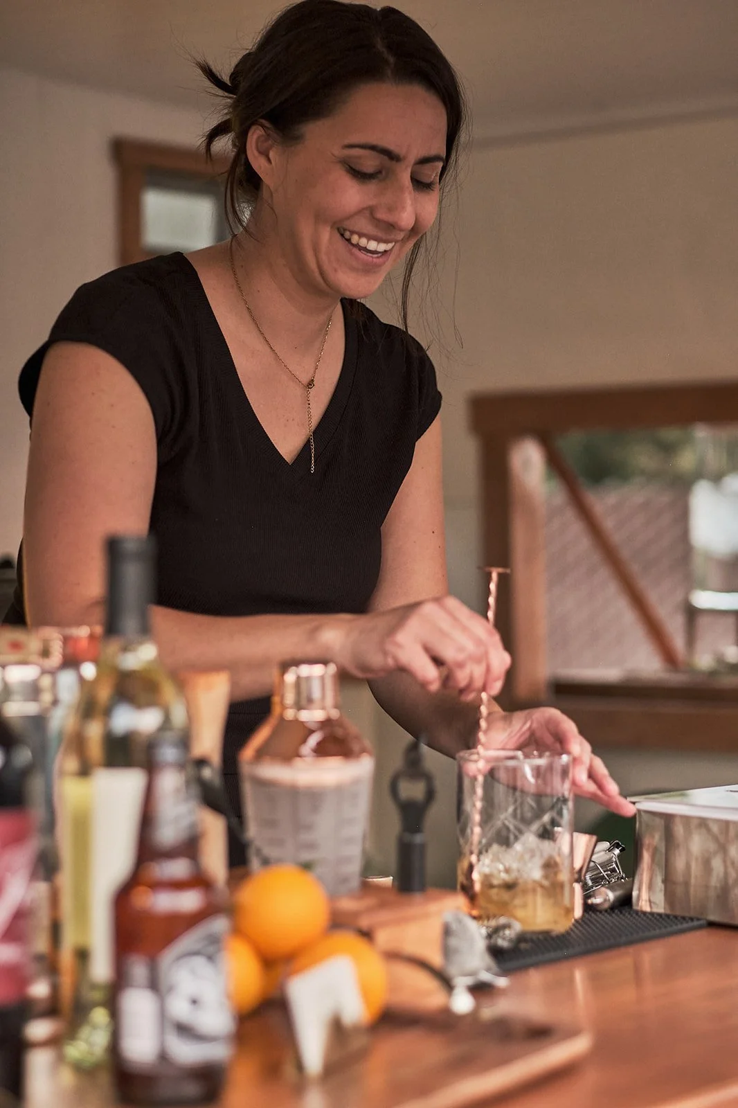 A woman with dark hair smiling and preparing a drink in a glass jar on a wooden counter. Towing Temptations Mobile Bar Trailer Fallbrook, ca