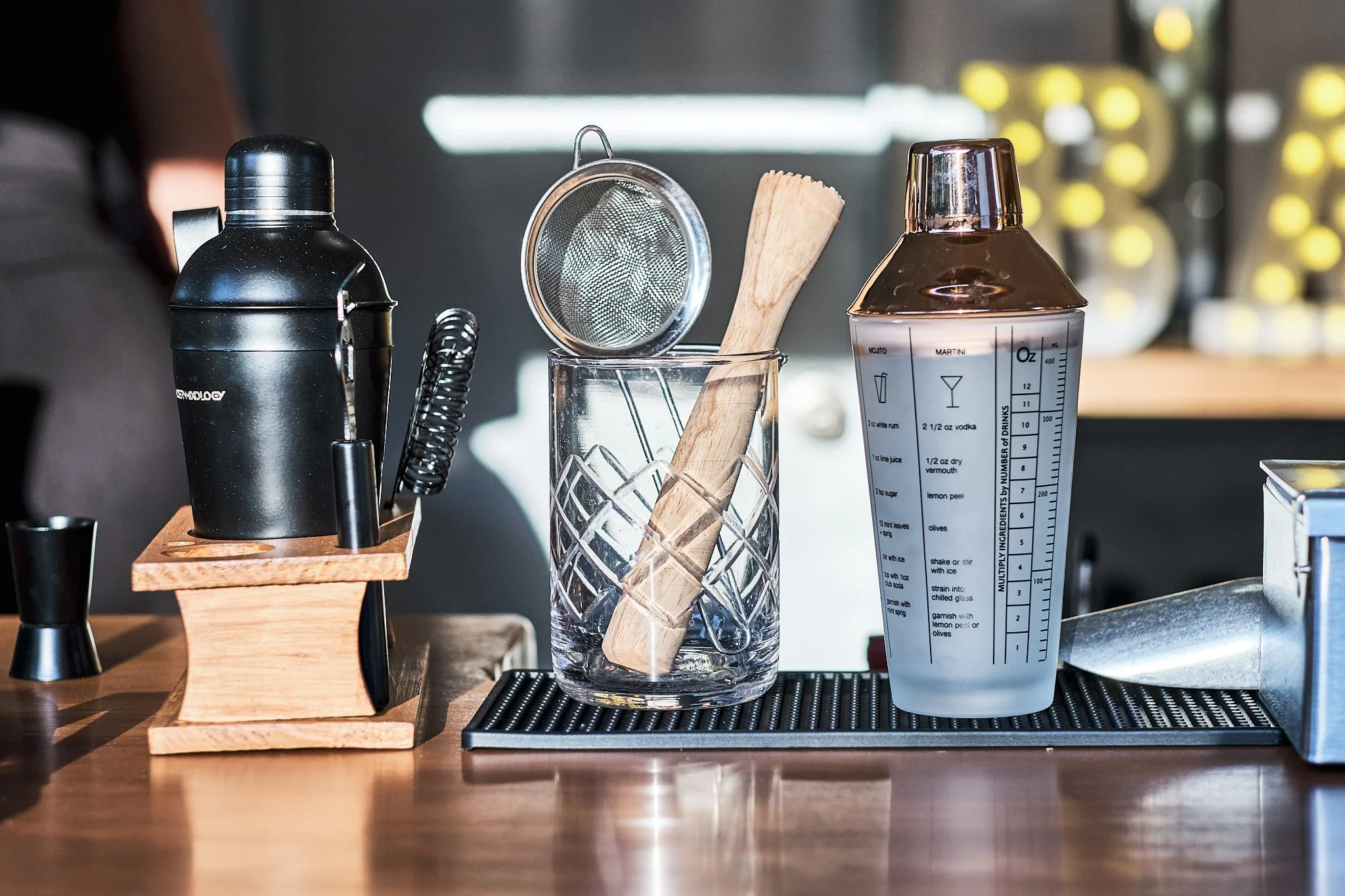 Bar tools and drink ingredients on a countertop, including a cocktail shaker, strainer, muddler, and a measuring bottle with cocktail recipes. Towing Temptations Mobile Bar Trailer