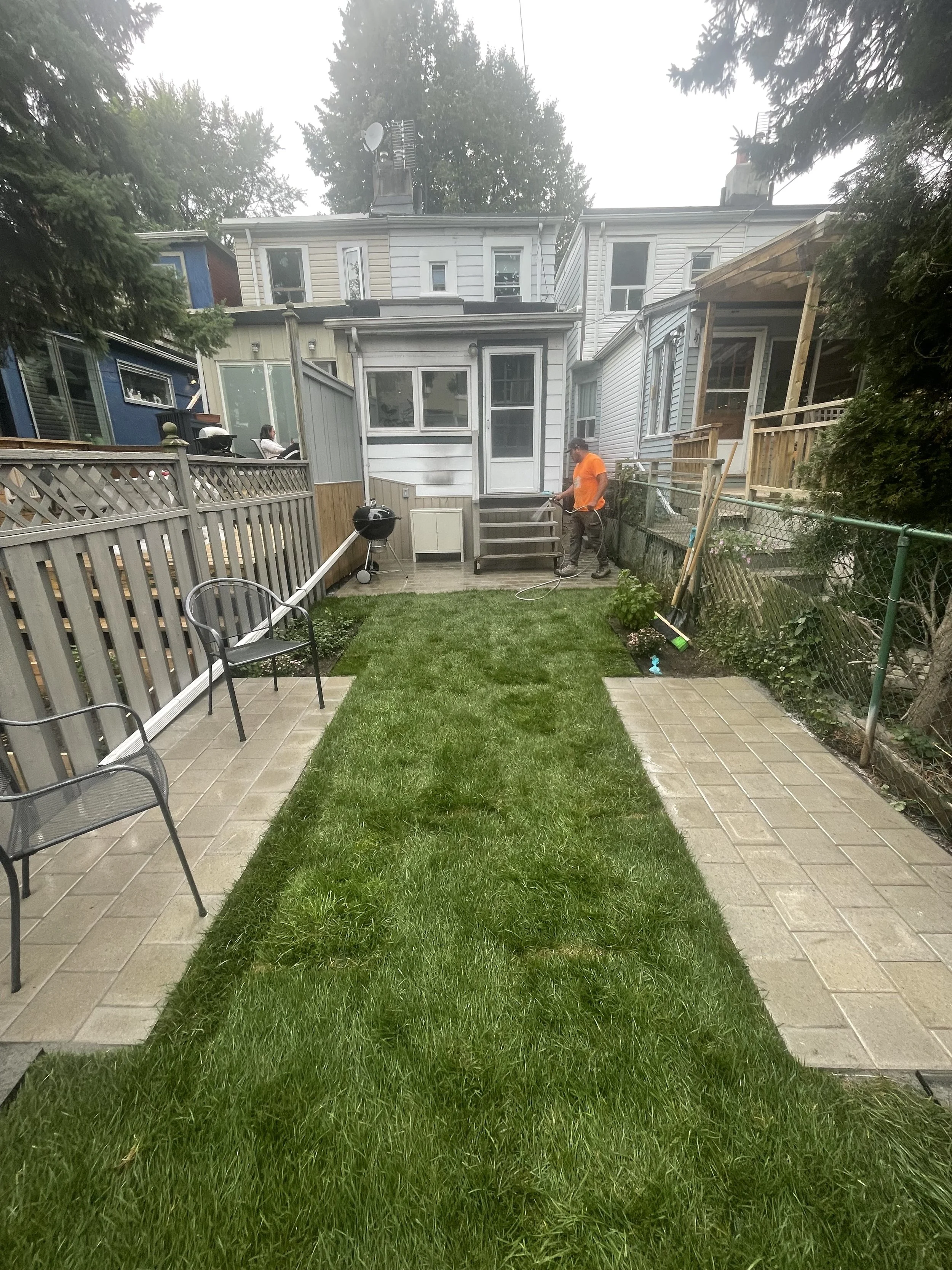 View of a small backyard with freshly laid grass, patio stones, and a person in an orange shirt holding a hose near the house. There is a fence, outdoor chairs, and a barbecue grill. Houses and trees are visible in the background.
