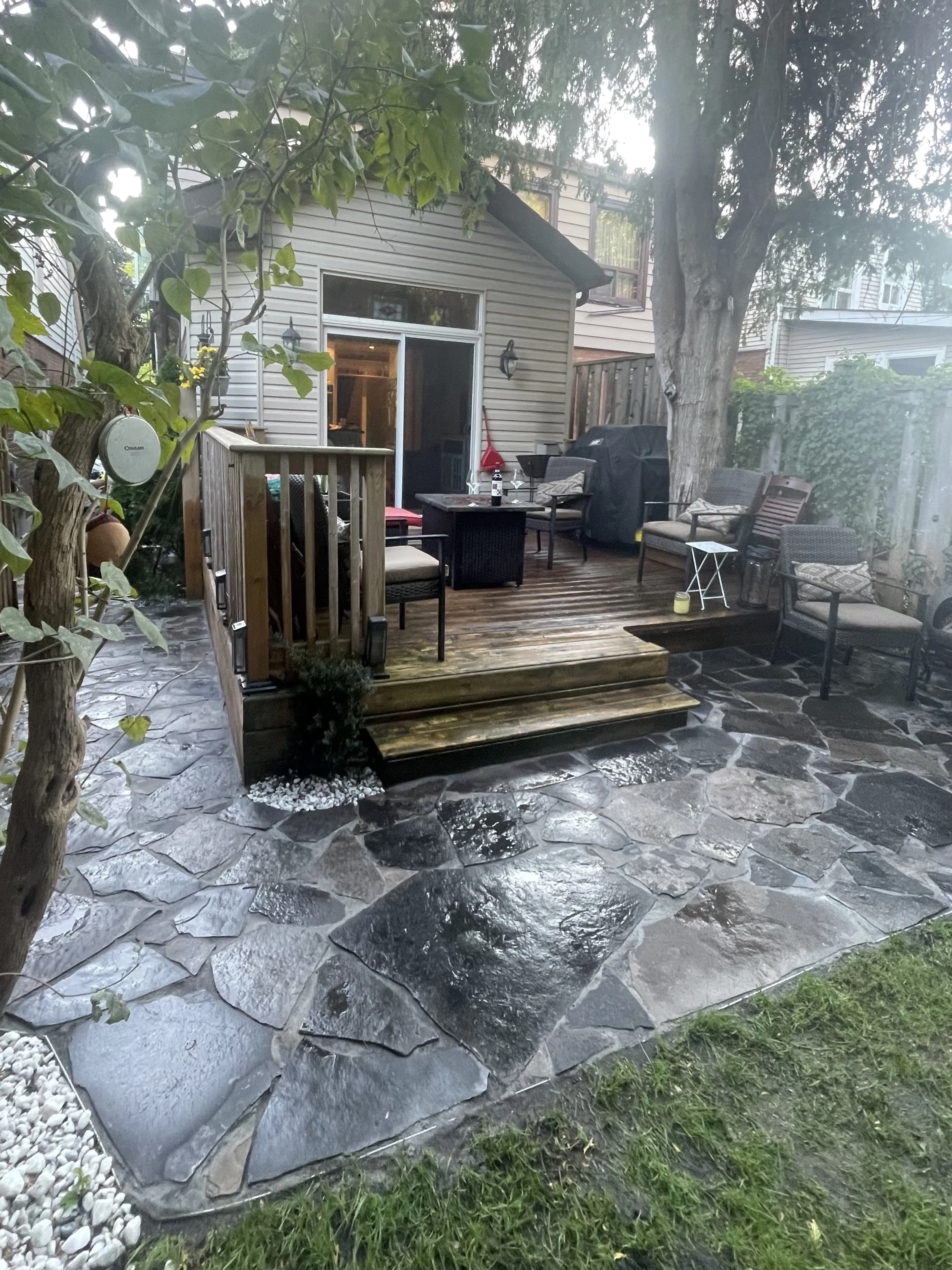 Backyard patio with stone tiles, wooden deck, outdoor furniture, and a large tree. A small house with sliding glass doors and surrounding greenery is in the background.