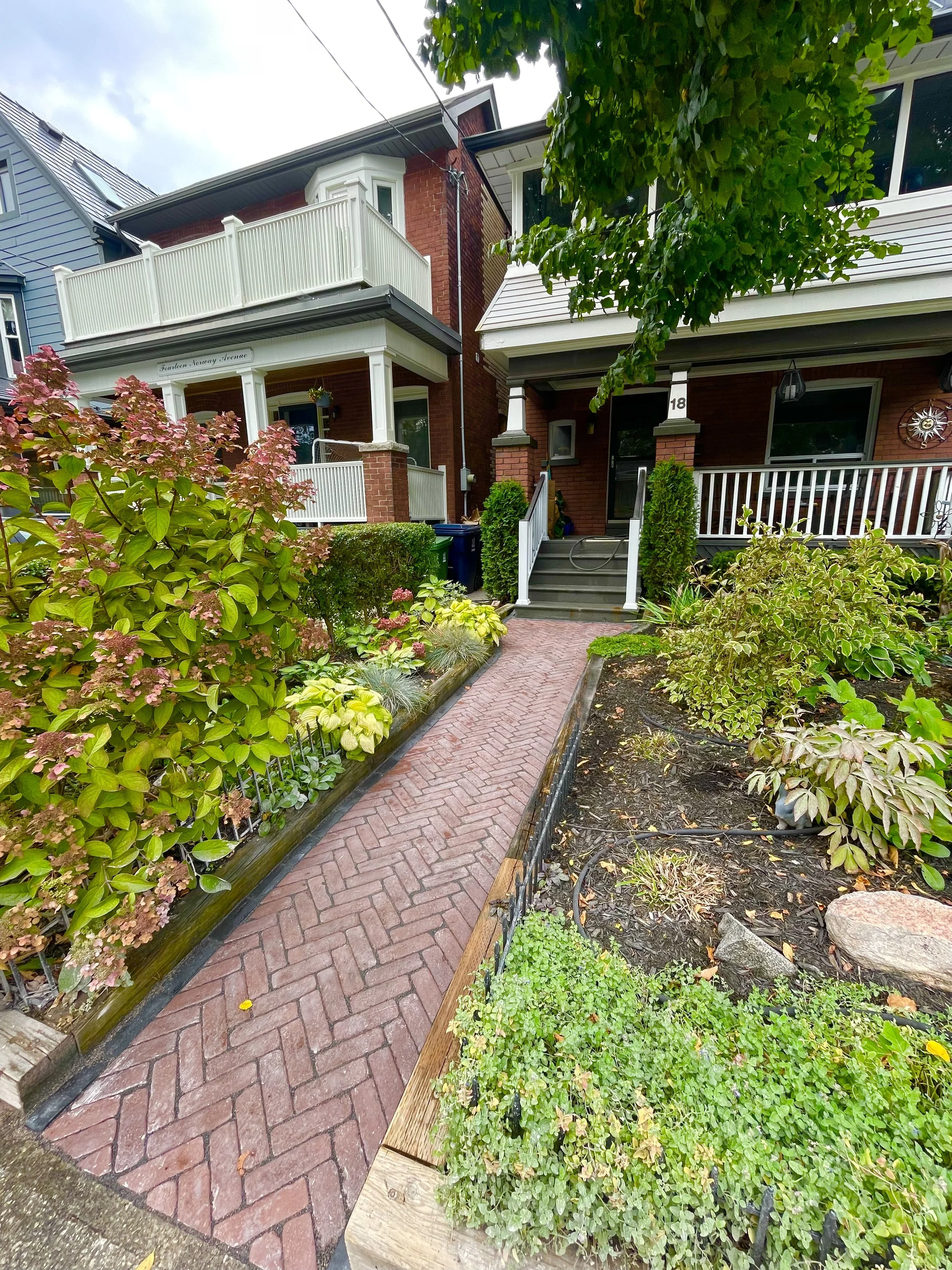 Brick pathway leading to a porch of a brick house with white trim, surrounded by lush greenery and plants.