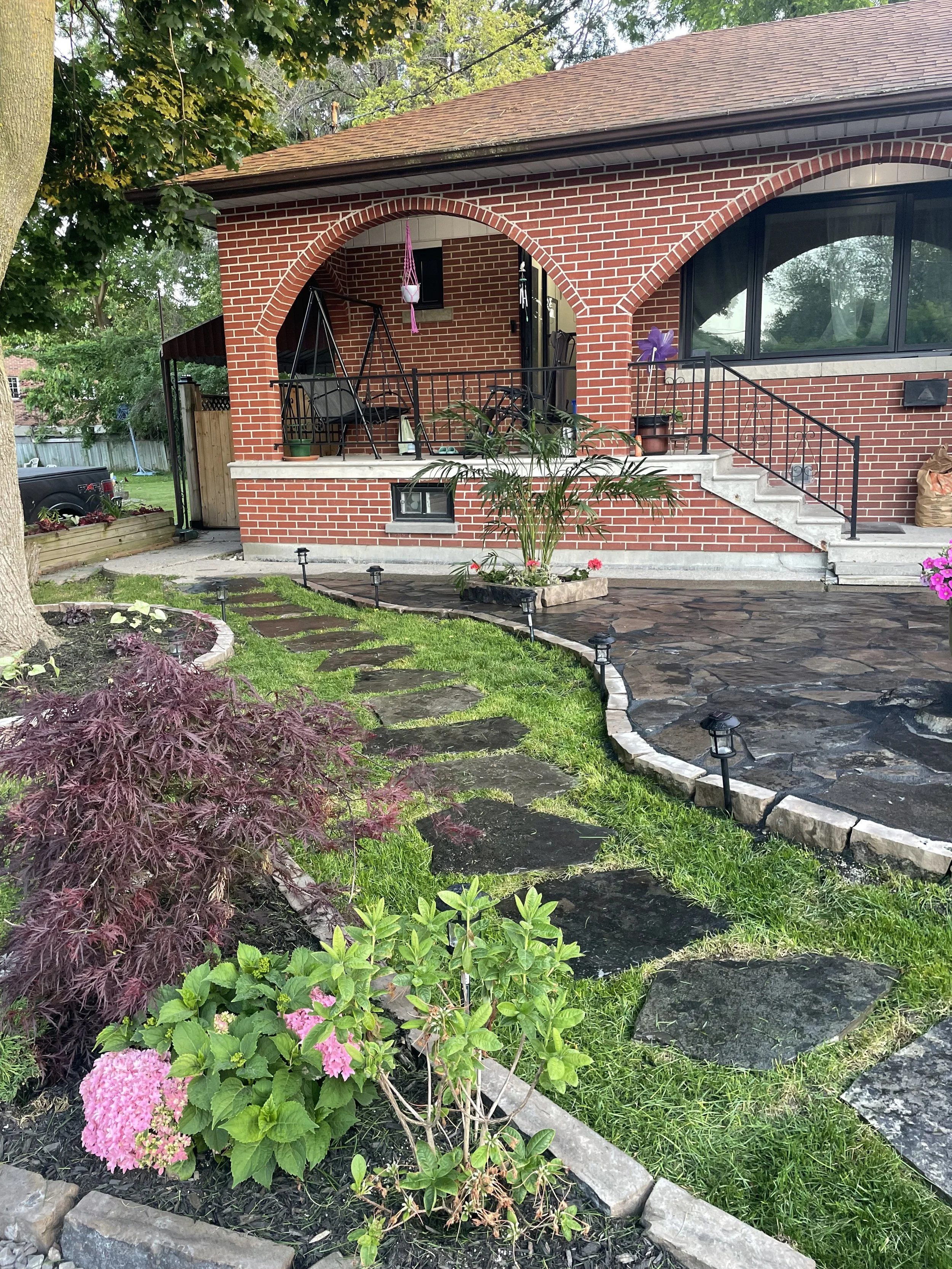 A cozy brick house with arched windows and a front porch swing surrounded by a well-maintained garden. The garden features stone pathways, lush green grass, vibrant hydrangeas, and other plants. The porch is accessible by stairs and lined with potted plants.