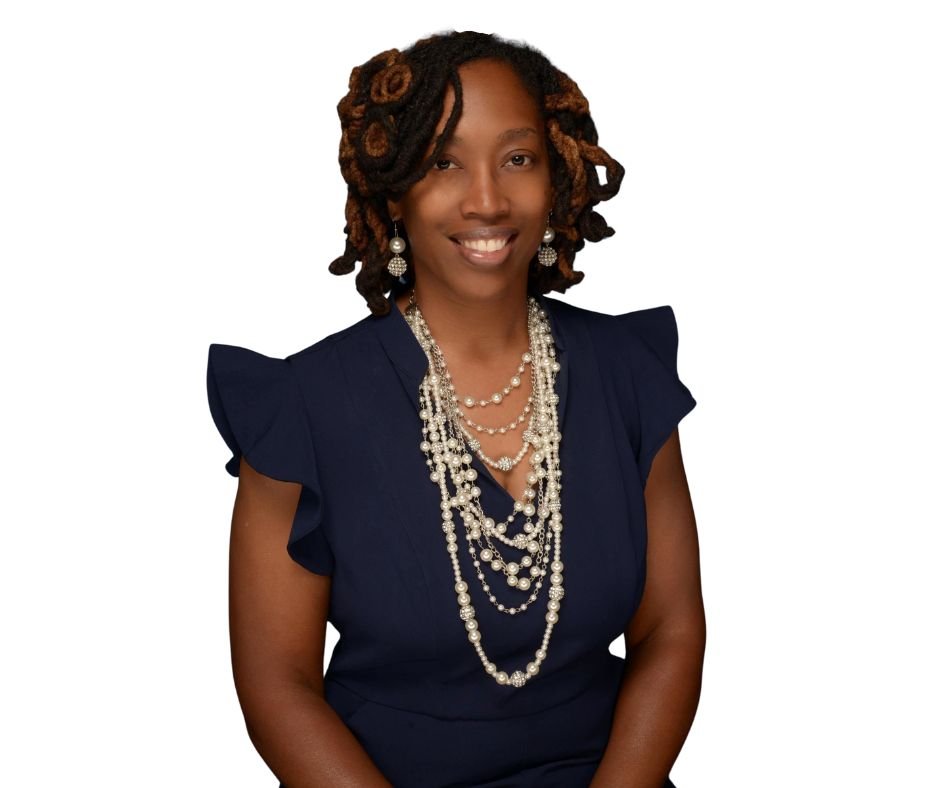 A smiling woman with dark skin and shoulder-length dreadlocks, wearing a navy blue dress with ruffled shoulders and layered pearl necklaces, standing against a white background.