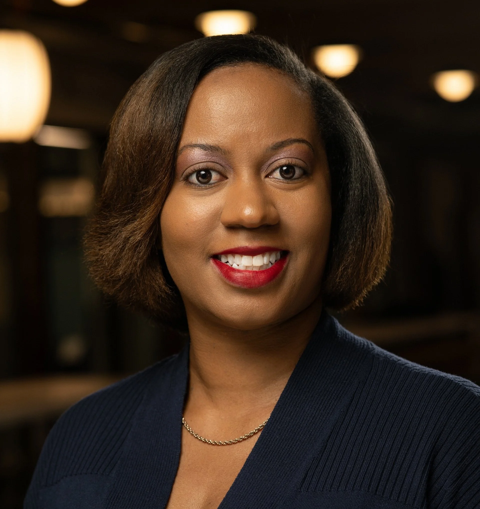 A professional woman with shoulder-length brown hair, smiling, wearing a navy blazer, gold necklace, and red lipstick, in a warmly lit indoor setting.