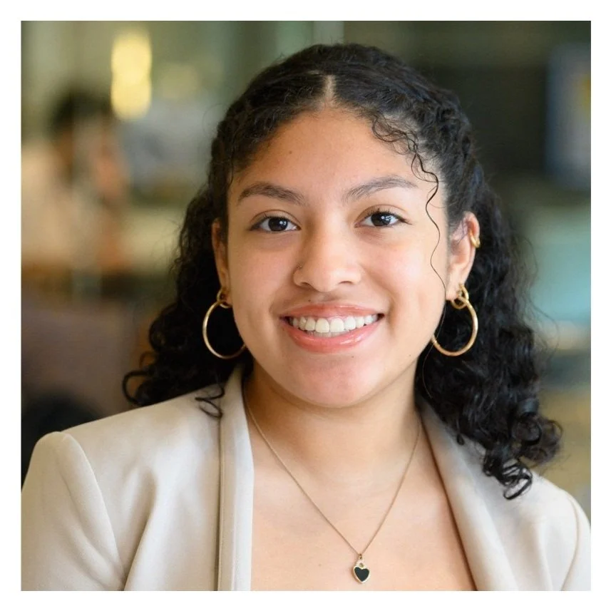 A young woman with curly dark hair, wearing gold hoop earrings, a beige blazer, and a necklace with a heart-shaped pendant, smiling at the camera with a blurred indoor background.