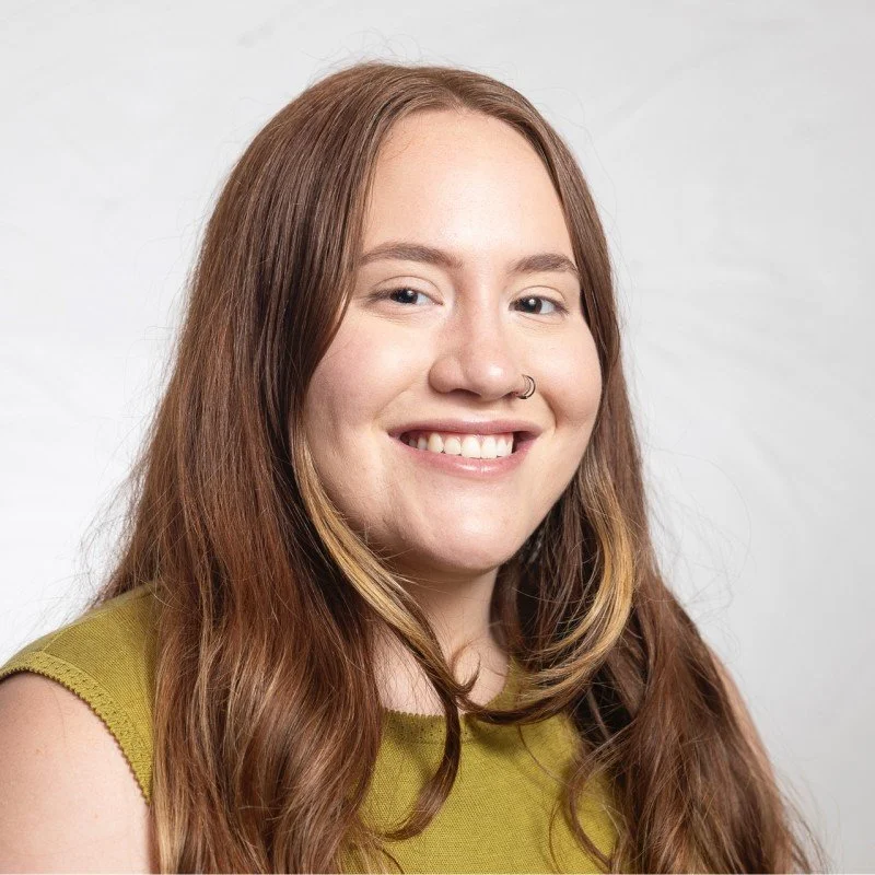 A young woman with long red hair, smiling, wearing a nose ring and a sleeveless green top, against a plain light background.