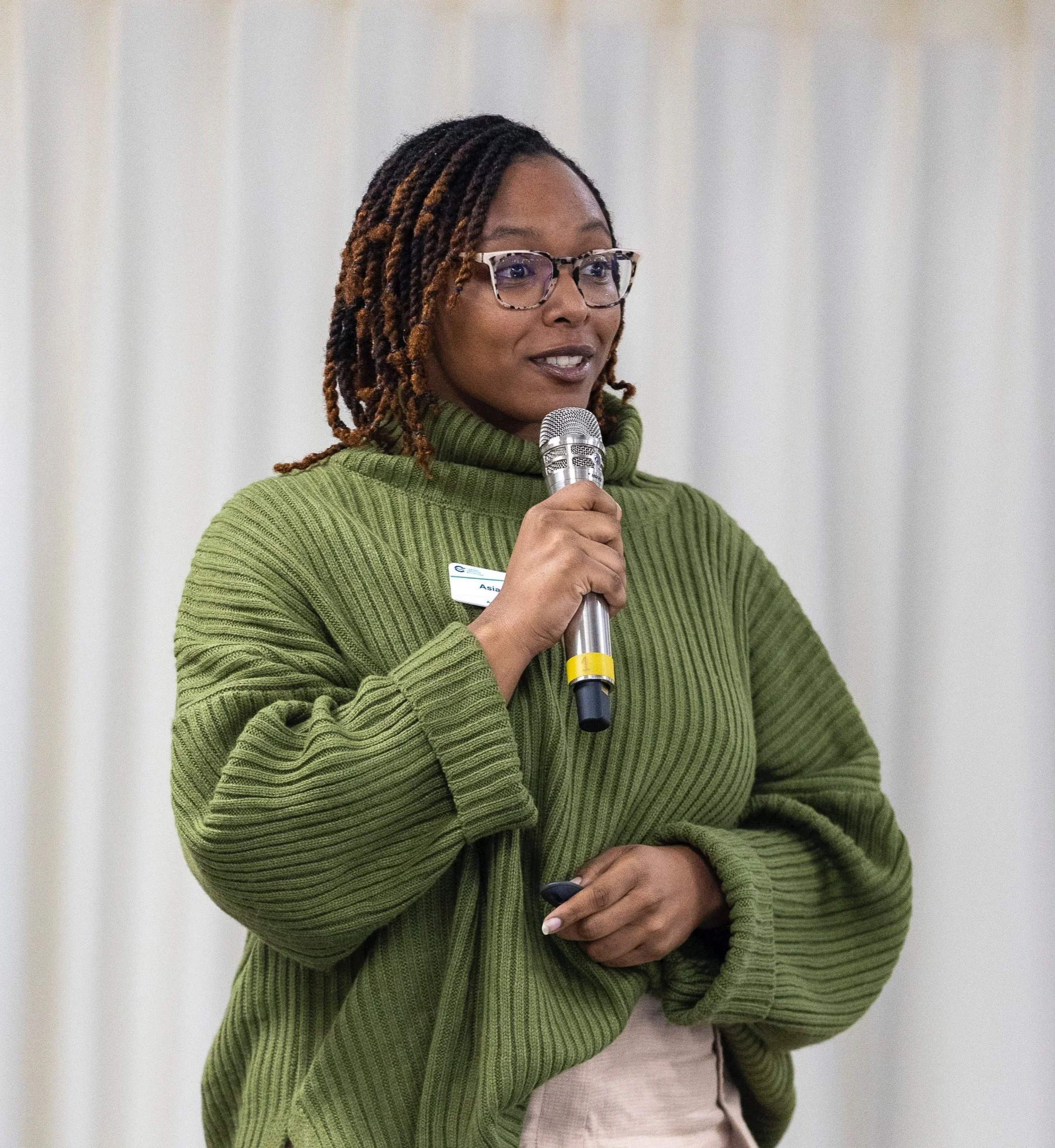 A woman with glasses and dreadlocks wearing a green sweater, speaking into a microphone at an indoor event.