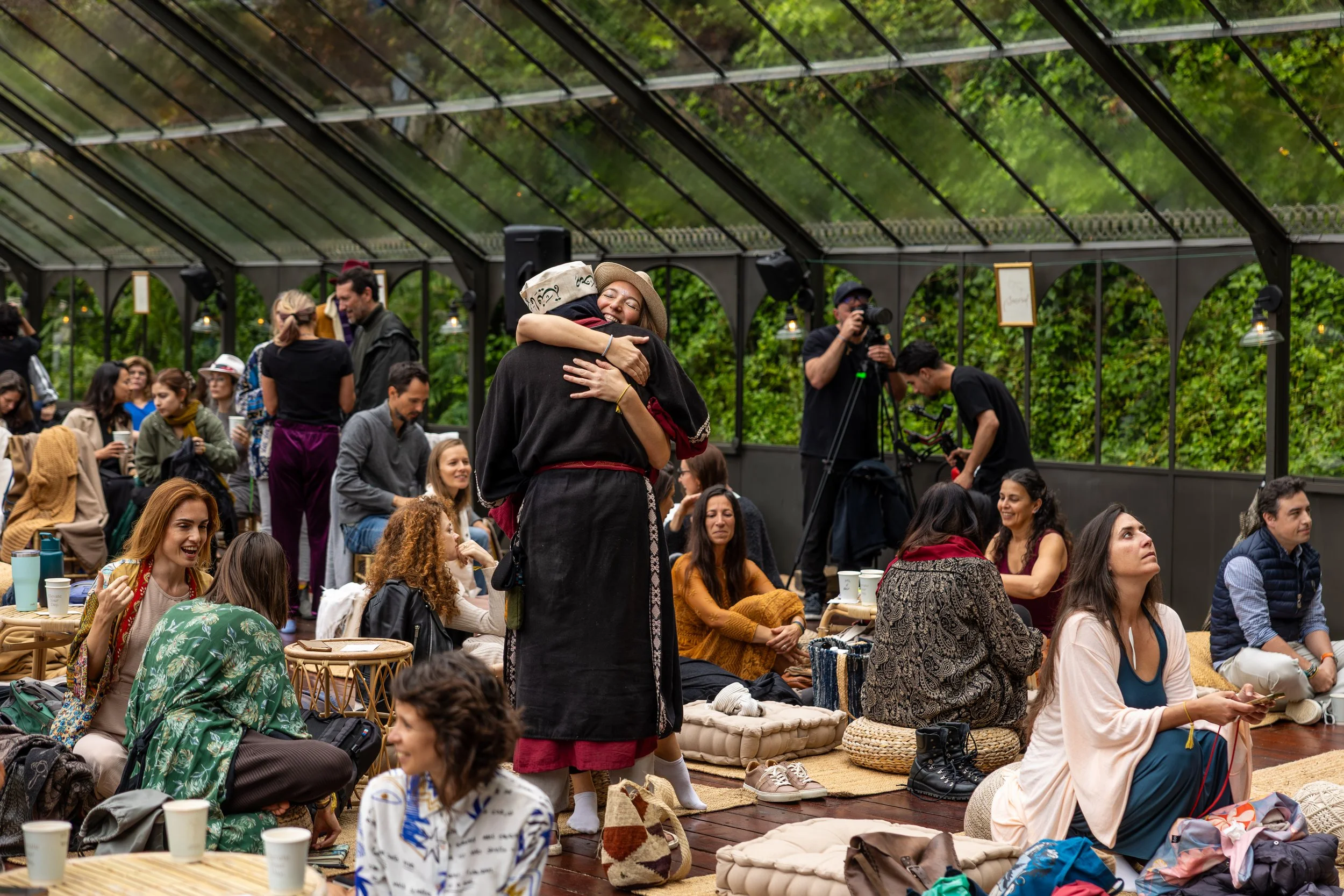 People gathered in a greenhouse, some sitting on cushions and others standing, with a woman and man hugging in the center. There are photographers and people using phones in the background at the FiVth Experience in Portugal.