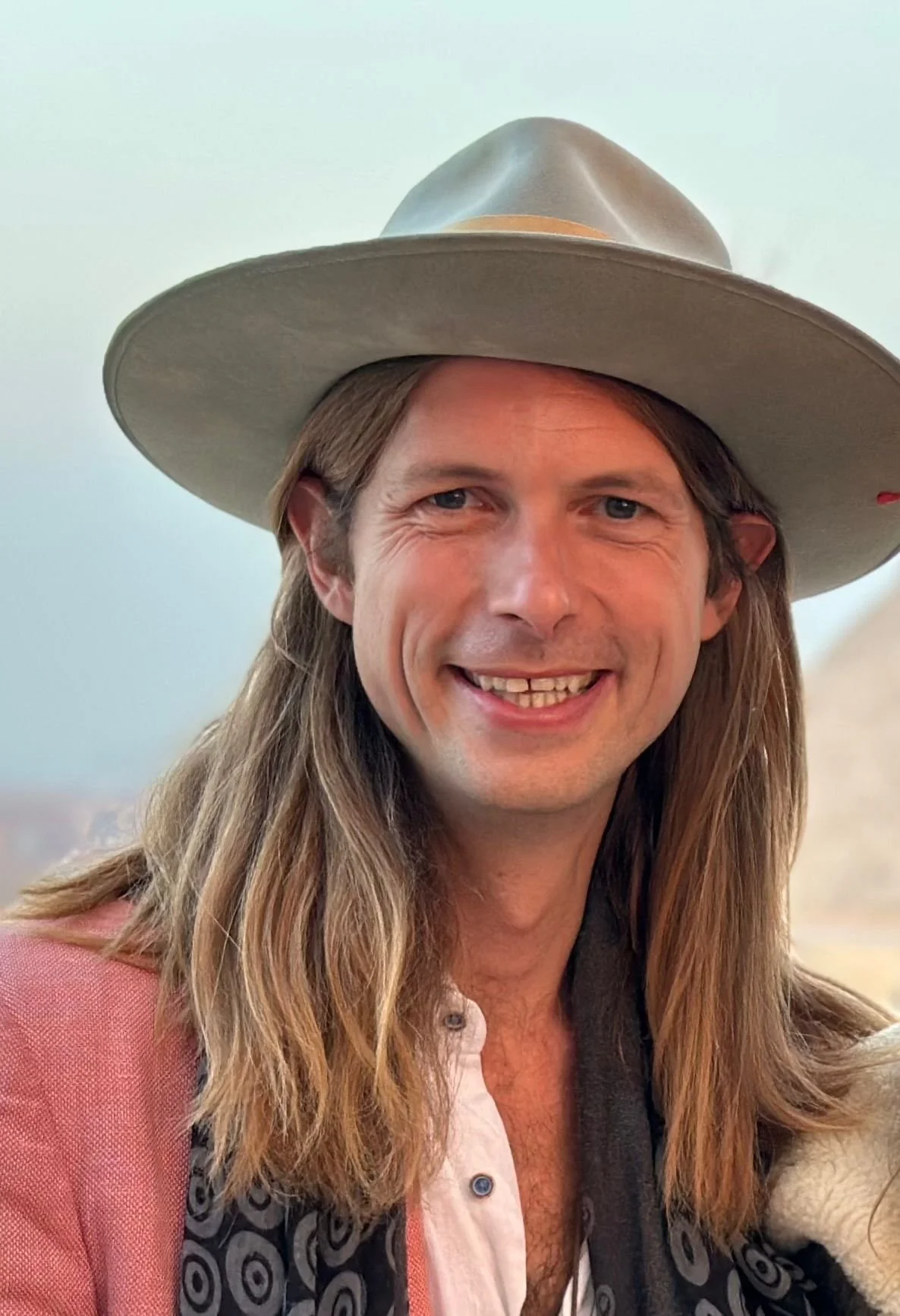 A man with long hair, wearing a large wide-brimmed hat, smiling, with a light-colored shirt and a dark scarf or jacket.