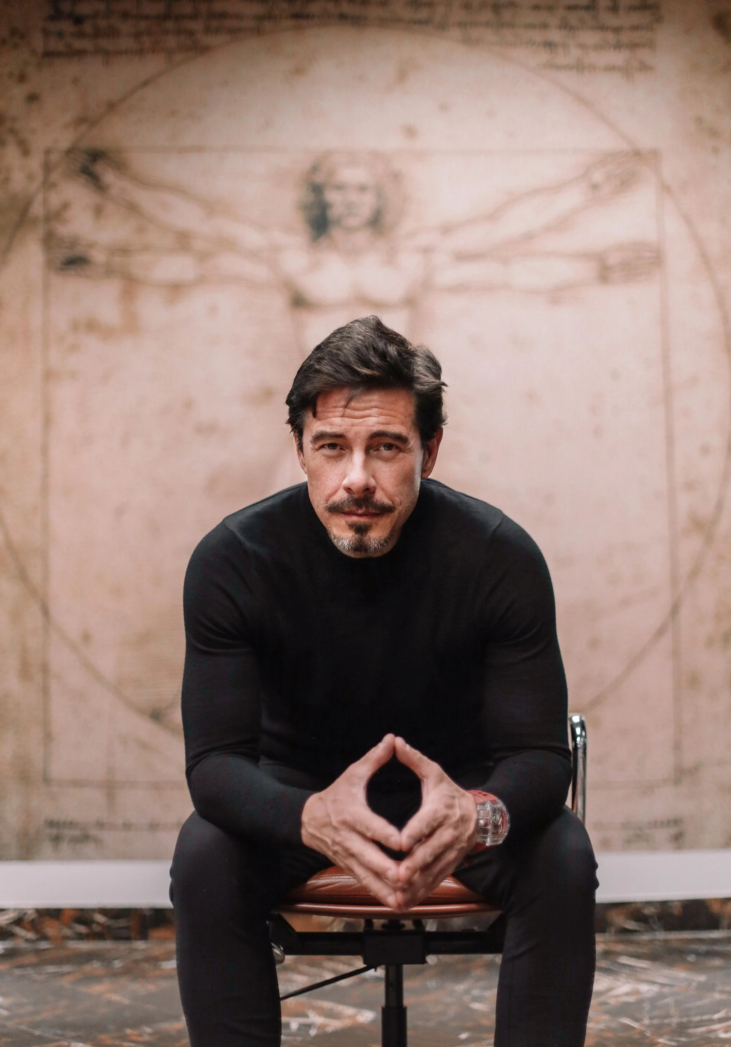 Robert Edward Grant sitting on a chair with hands clasped, in front of a background featuring Leonardo da Vinci's Vitruvian Man drawing.
