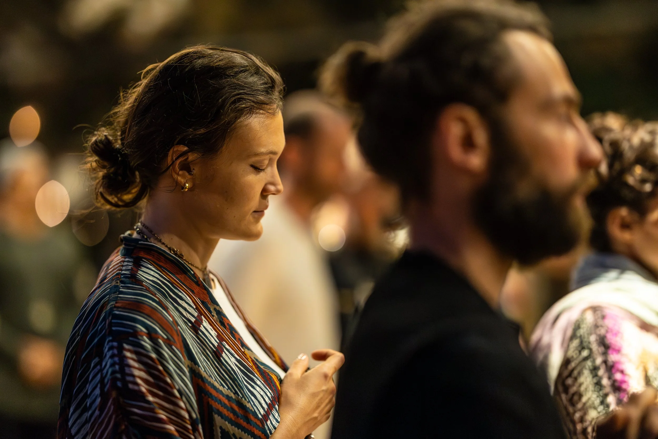 A woman with dark hair tied in a bun, wearing a patterned shirt, and gold earrings, standing with her eyes closed and hands clasped, in a prayer or meditative posture among a group of people in a dimly lit setting at the FiVth Experience in Portugal.