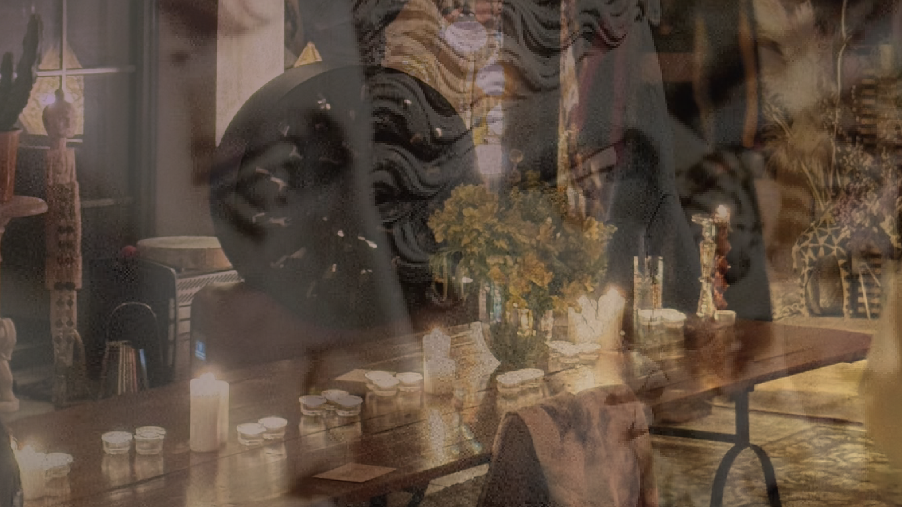 A table with candles, vases, and a floral centerpiece in a room decorated with African masks, figurines, and textiles, possibly inside a house or gallery.