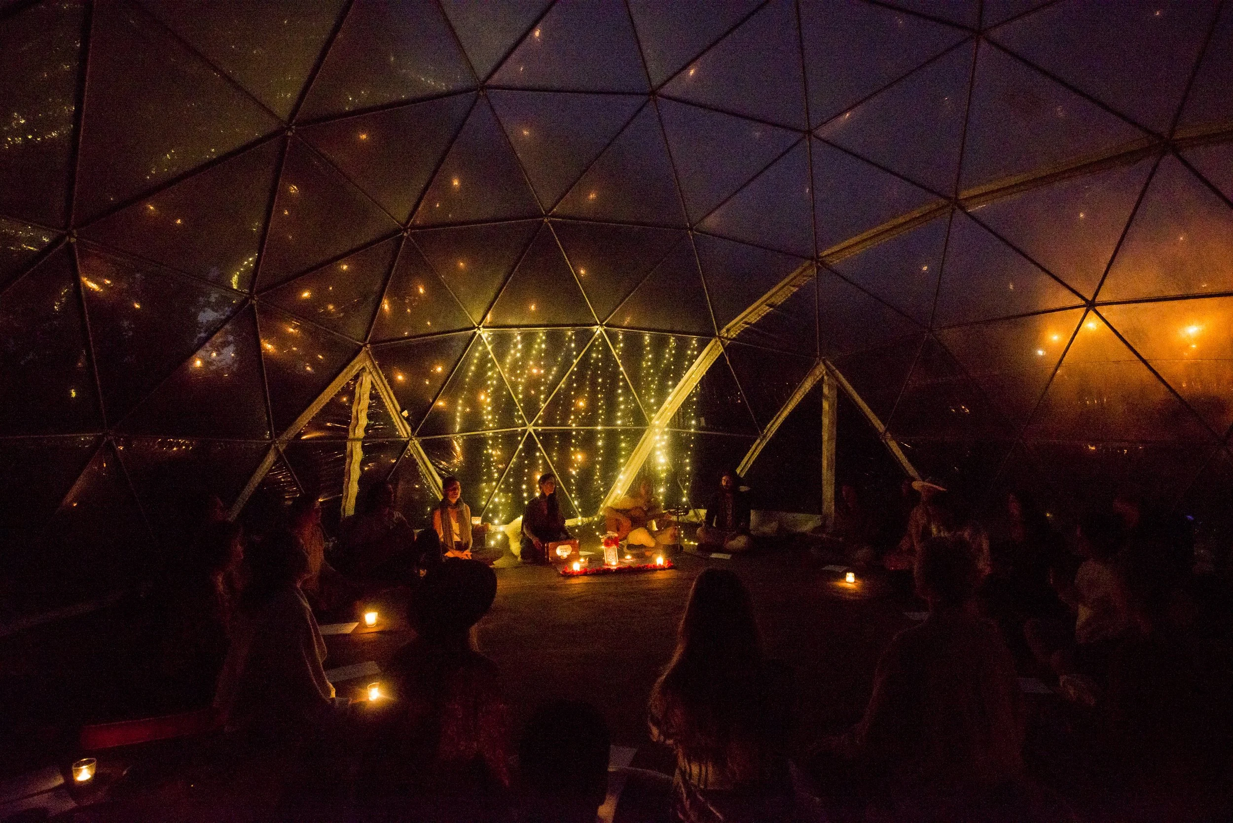 People sitting inside a geodesic dome at night, illuminated by string lights and candles, attending a music or meditation session at a conscious event in Ericeira.